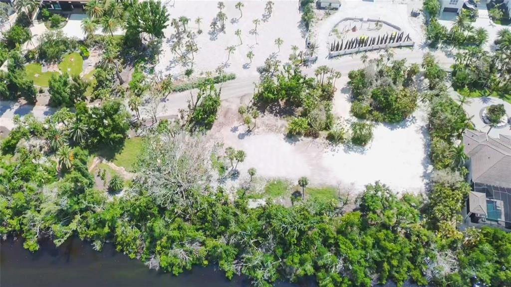 An aerial view shows a vacant, sandy waterfront lot surrounded by lush trees and nearby homes — 7825 SANDERLING ROAD, SARASOTA, FL