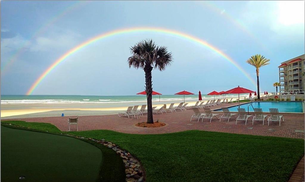 A double rainbow arches over a beachside pool deck with lounge chairs and palm trees — 5501 ATLANTIC UNIT 107, NEW SMYRNA BEACH, FL