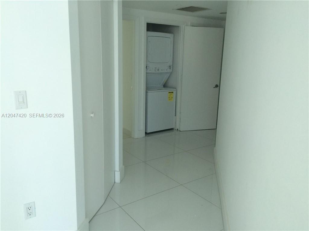 A white hallway with tile flooring leads to a closet containing a stacked washer-dryer unit — 55 SE 6th St # 2807, Miami FL 33131, MIAMI, FL