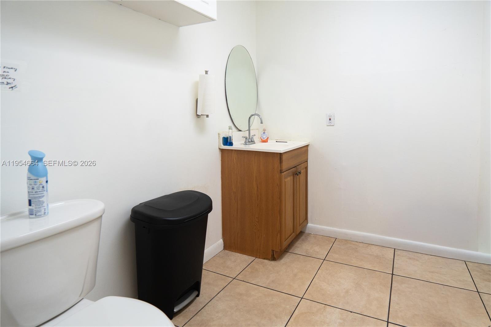 A bathroom features a wooden vanity, a small oval mirror, and a black trash can — 12253 S Dixie Hwy, Pinecrest FL 33156, PINECREST, FL