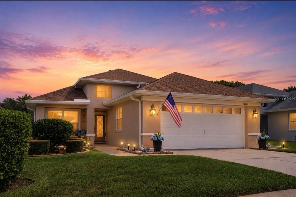A tan single-story house with a two-car garage and an American flag at sunset — 14439 MIDDLE FAIRWAY DRIVE, SPRING HILL, FL