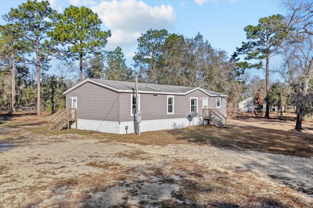 A gray manufactured home with white skirting sits on a dirt lot among pine trees — 1910 NE 155TH COURT, WILLISTON, FL