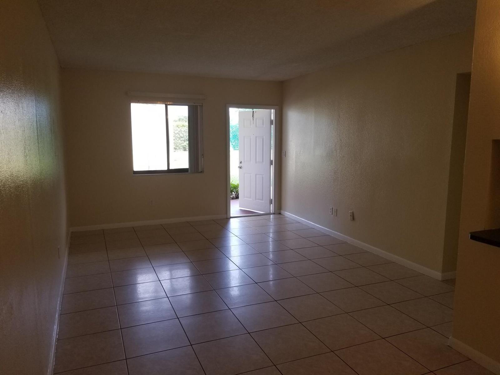 An empty living room features beige walls, light-colored tile flooring, a window, and an open door — 6023 10th Avenue N # 114, FL 33463, FL