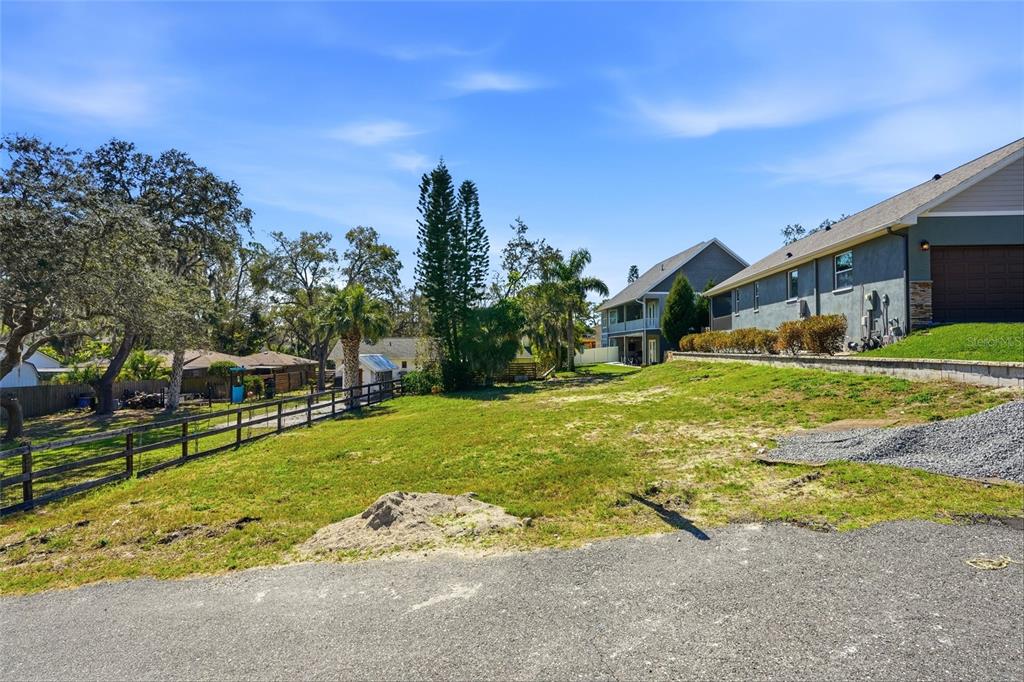 A grassy vacant lot sits next to a house with a gravel driveway and fence — 1604 BUTLER COURT, TARPON SPRINGS, FL