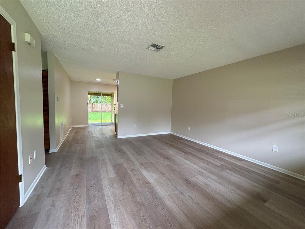 An empty living room with light-colored walls and wood-look flooring leads to a sliding glass door — 20015 SUNCREST DRIVE, BROOKSVILLE, FL