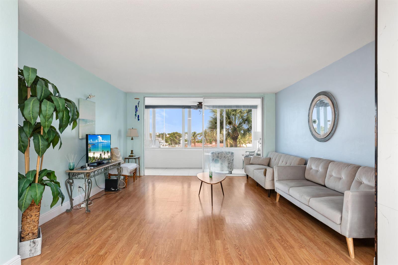 A living room features light blue walls, wood flooring, a grey sofa, and a balcony view — 401 N Riverside Drive # 406, Pompano Beach FL 33062, POMPANO BEACH, FL