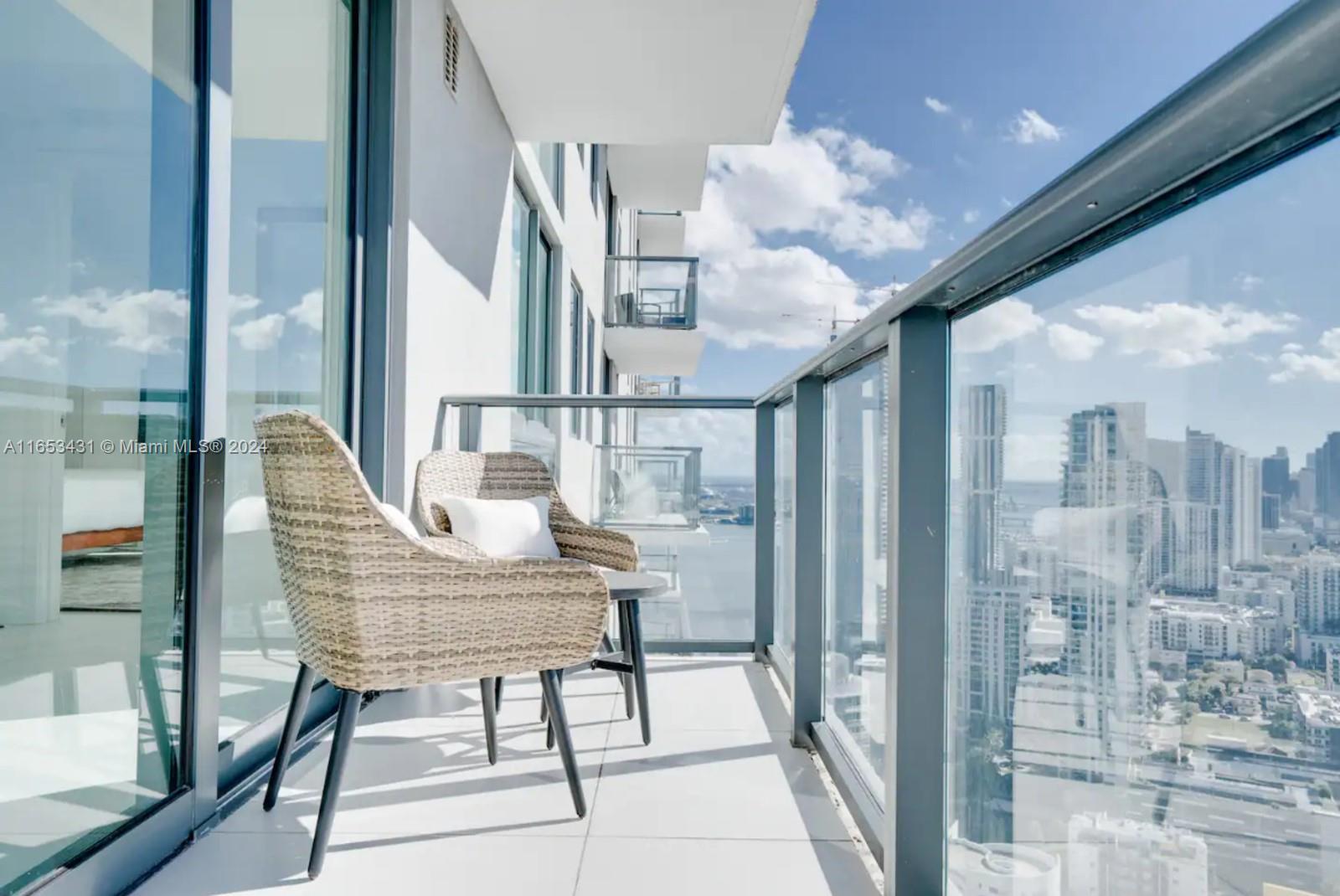 A high-rise balcony features two wicker chairs, a small table, and glass railings overlooking city buildings — 501 NE 31st St # 3709, Miami FL 33137, MIAMI, FL