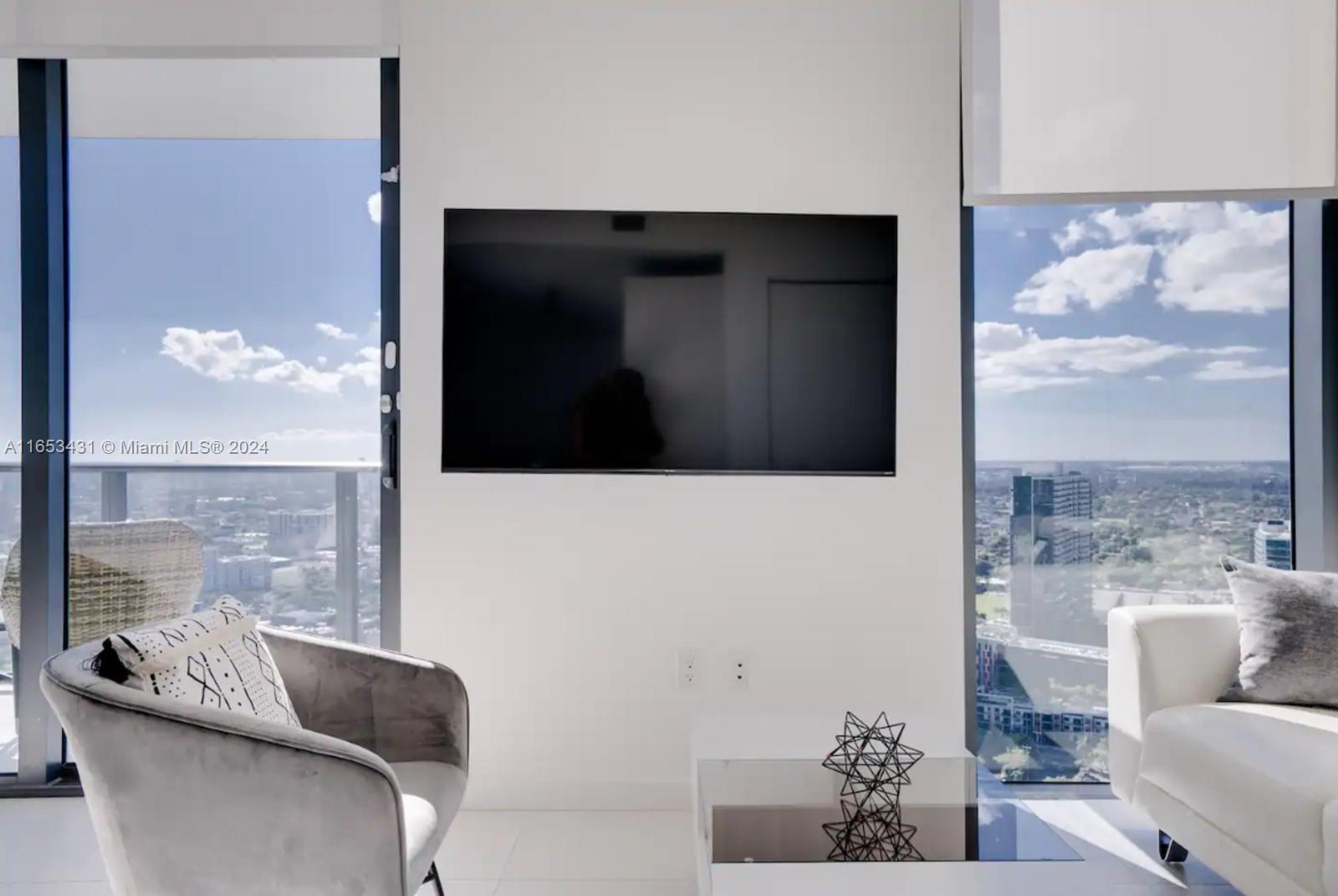 A modern living room features a wall-mounted television between two floor-to-ceiling windows overlooking a city — 501 NE 31st St # 3709, Miami FL 33137, MIAMI, FL