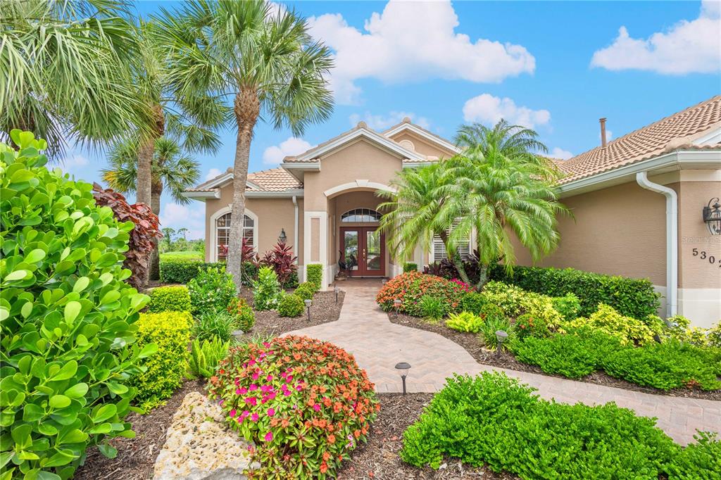 A paved walkway leads to the front entrance of a tan house with palm trees — 5302 WHITE IBIS CT., NORTH PORT, FL