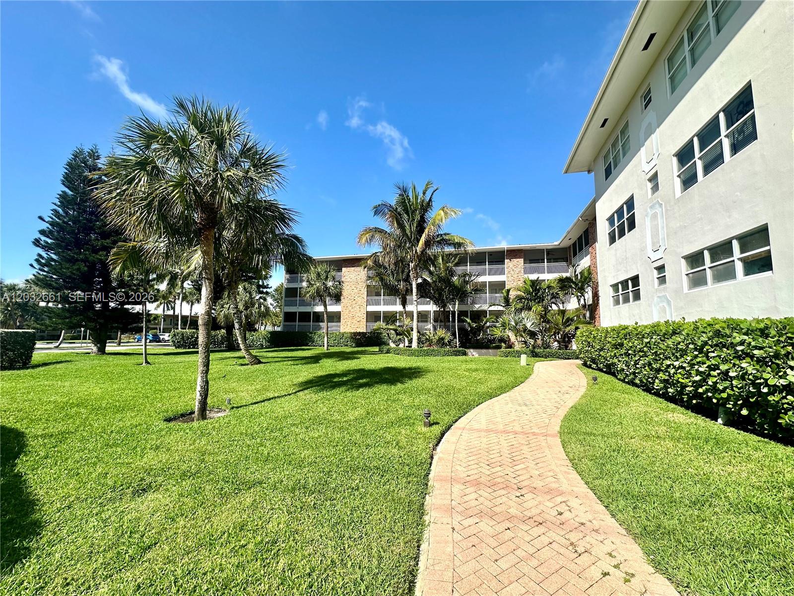 A brick walkway leads through a grassy courtyard toward a multi-story apartment building with palms — 2501 S Ocean Blvd # 1040, Boca Raton FL 33432, BOCA RATON, FL