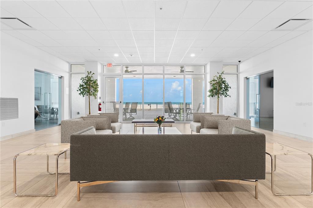 A modern lobby features gray sofas, marble-topped side tables, and floor-to-ceiling windows overlooking the ocean — 1700 BENJAMIN FRANKLIN DRIVE UNIT 10F, SARASOTA, FL