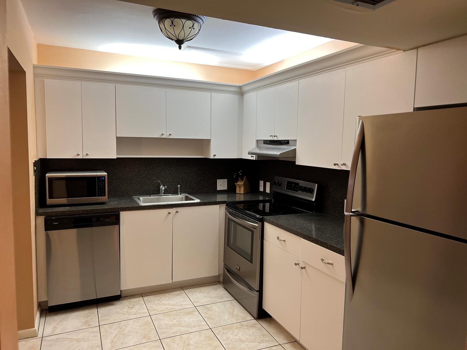 A kitchen with white cabinets, stainless steel appliances, dark countertops, and light tile flooring — 203 Suffolk, Boca Raton FL 33434, BOCA RATON, FL