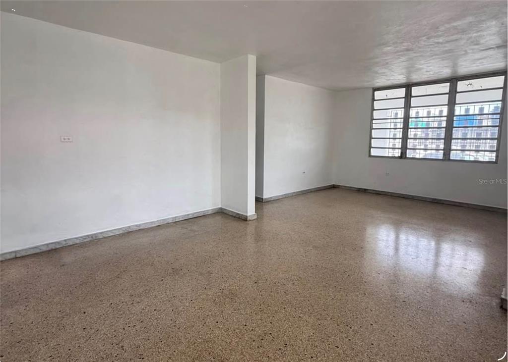 An empty room with white walls, terrazzo flooring, and large windows overlooking a city building — . ACACIA UNIT A37, CANOVANAS, PR