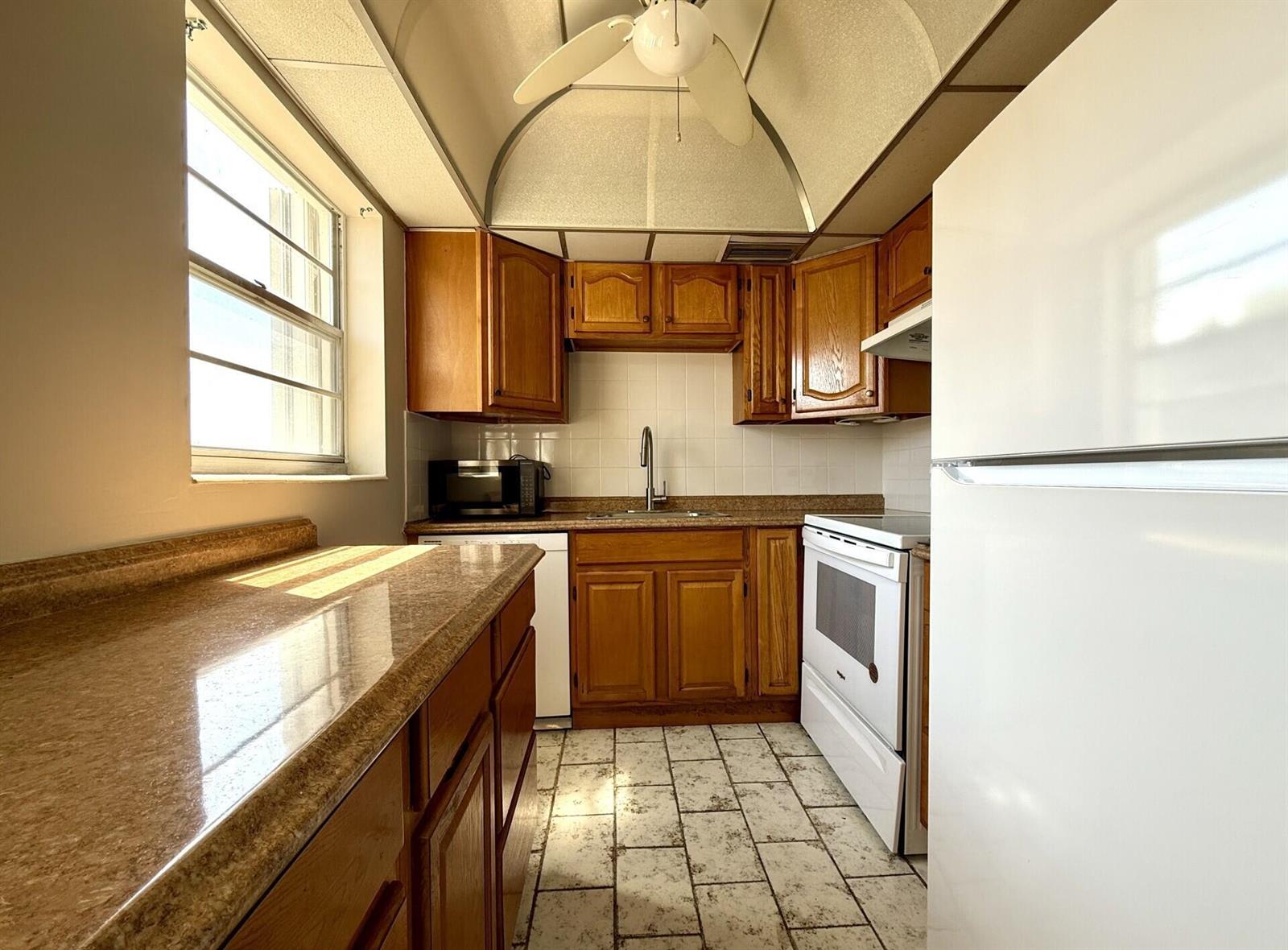 A galley kitchen features wood cabinets, laminate countertops, white appliances, and a vaulted ceiling — 93 Sheffield D # 93, West Palm Beach FL 33417, WEST PALM BEACH, FL