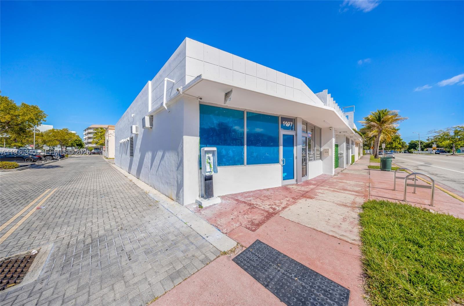 A white commercial building with a blue-tinted storefront stands beside a paved parking lot — 1975 71st St, Miami Beach FL 33141, MIAMI BEACH, FL