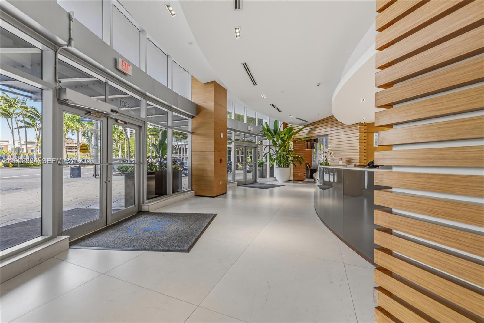 A modern building lobby features a curved reception desk, wood-slatted walls, and floor-to-ceiling windows — 3301 NE 1st Ave # H2011, Miami FL 33137, MIAMI, FL