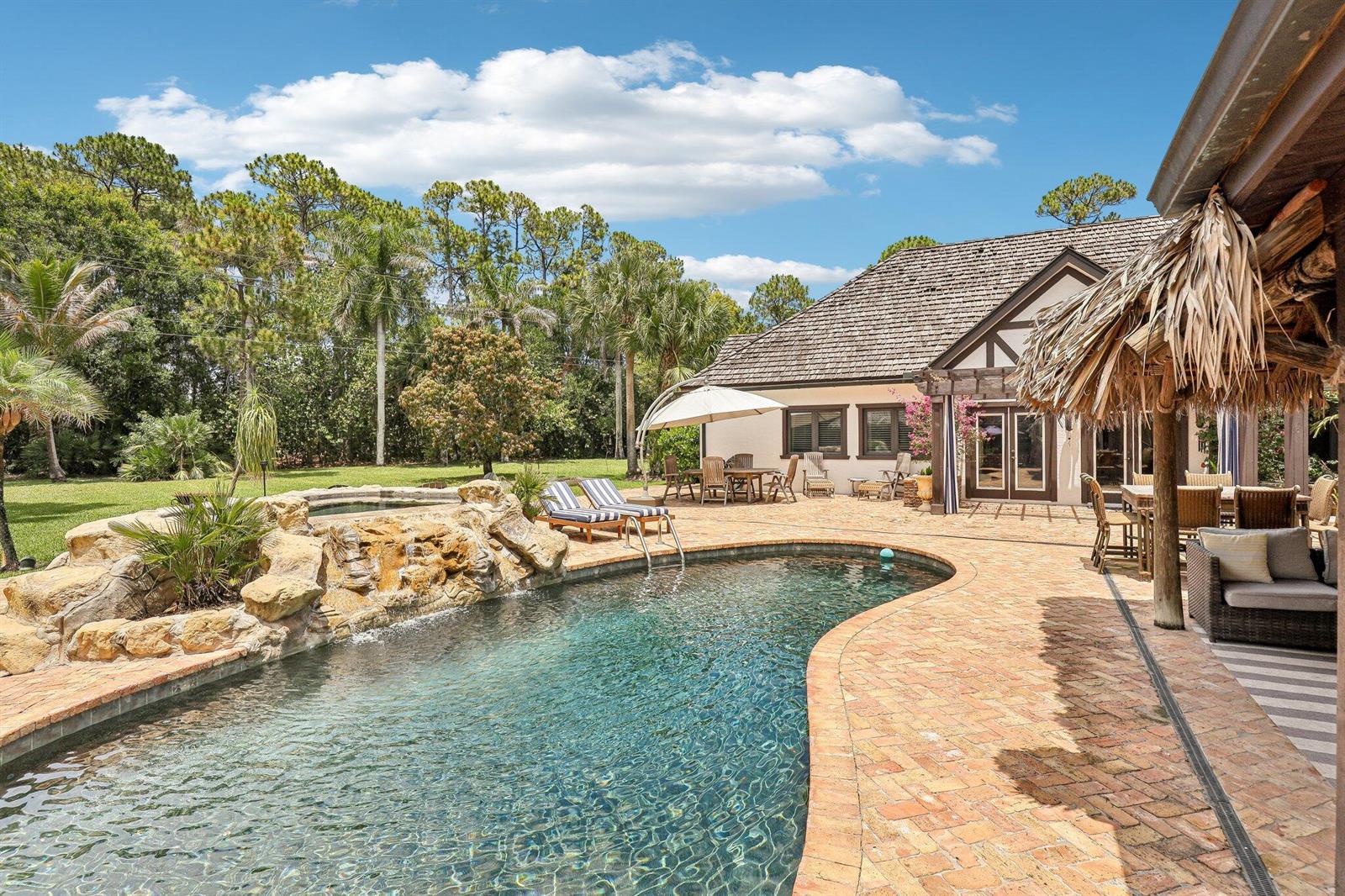 A backyard features a swimming pool with a rock waterfall, brick patio, and outdoor seating — 14900 Stirrup Lane, Wellington FL 33414, WELLINGTON, FL