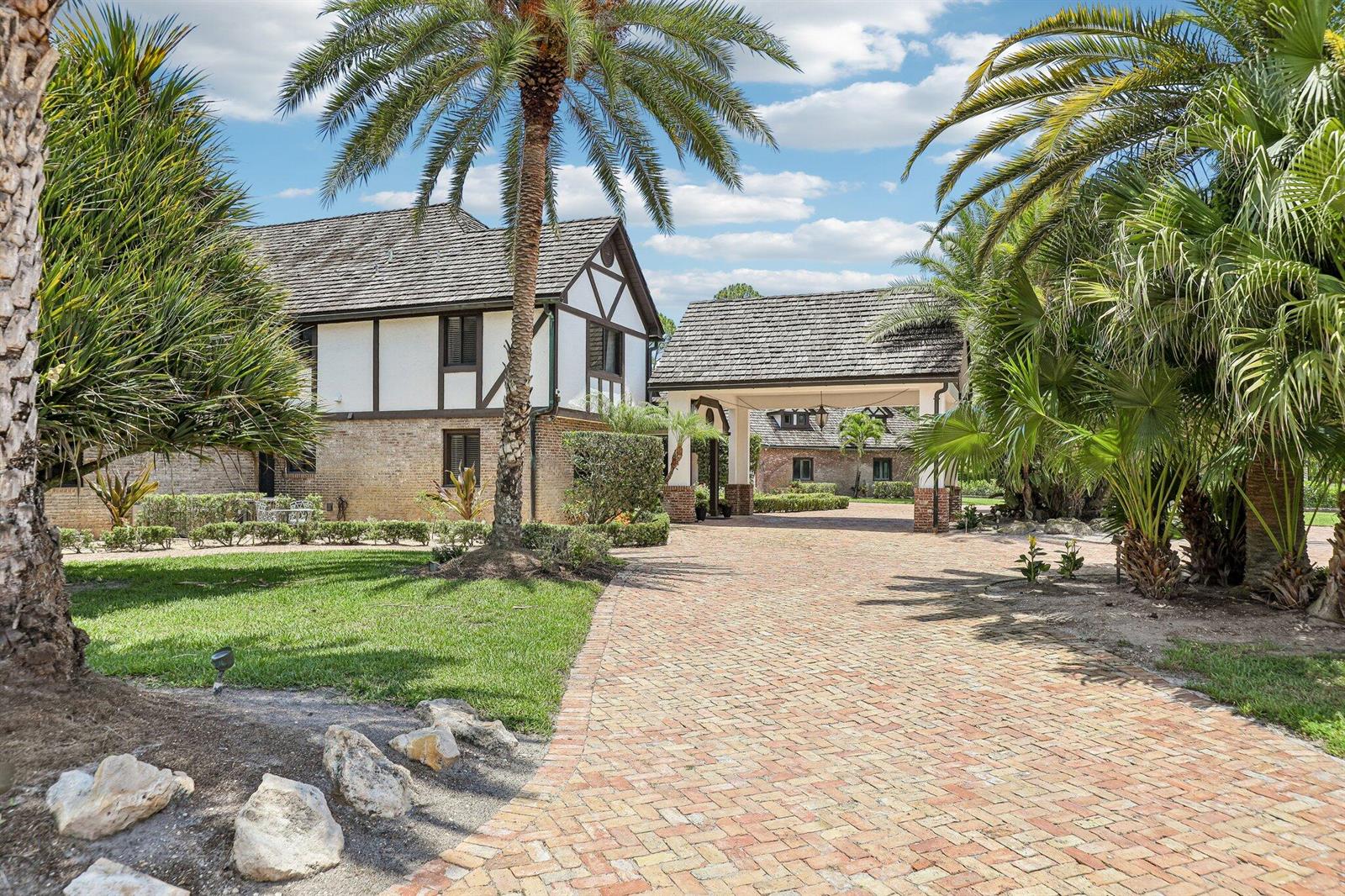 A brick driveway leads to a Tudor-style house with a covered porte-cochère and palm trees — 14900 Stirrup Lane, Wellington FL 33414, WELLINGTON, FL