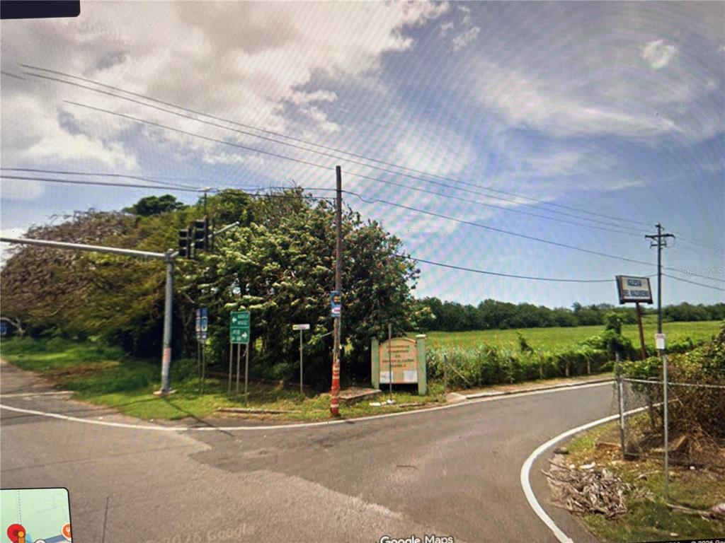 An outdoor view of a road intersection with a sign, traffic lights, and grassy fields — PR 2 KM. 115.5, AGUADILLA, PR