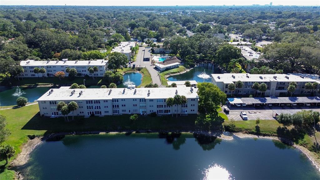 An aerial view shows apartment buildings surrounding a lake with fountains and parking areas — 936 VIRGINIA STREET UNIT 206, DUNEDIN, FL