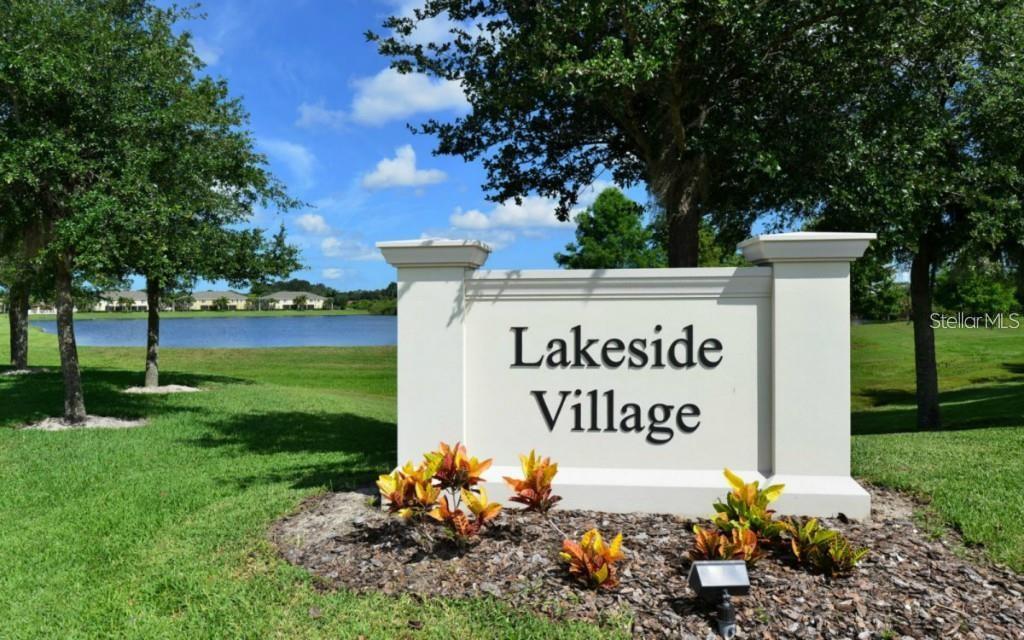 A white monument sign for Lakeside Village stands in a grassy area near a lake — 3909 45TH TERRACE W UNIT 106, BRADENTON, FL
