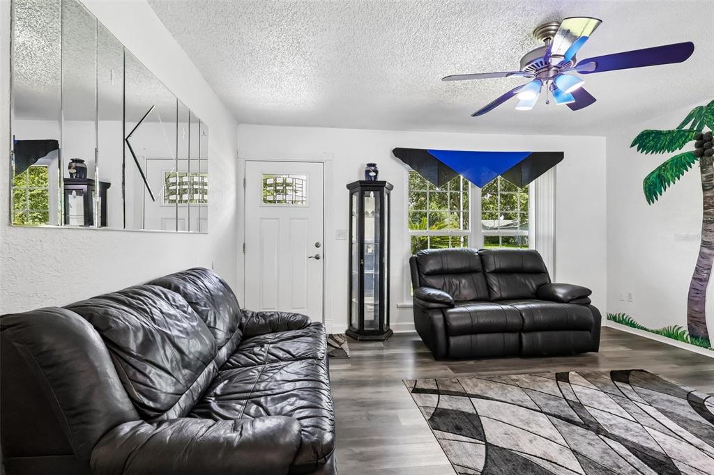 A living room features gray wood-look flooring, two black leather sofas, and a palm tree mural — 6513 NEWMAN CIRCLE W, LAKELAND, FL