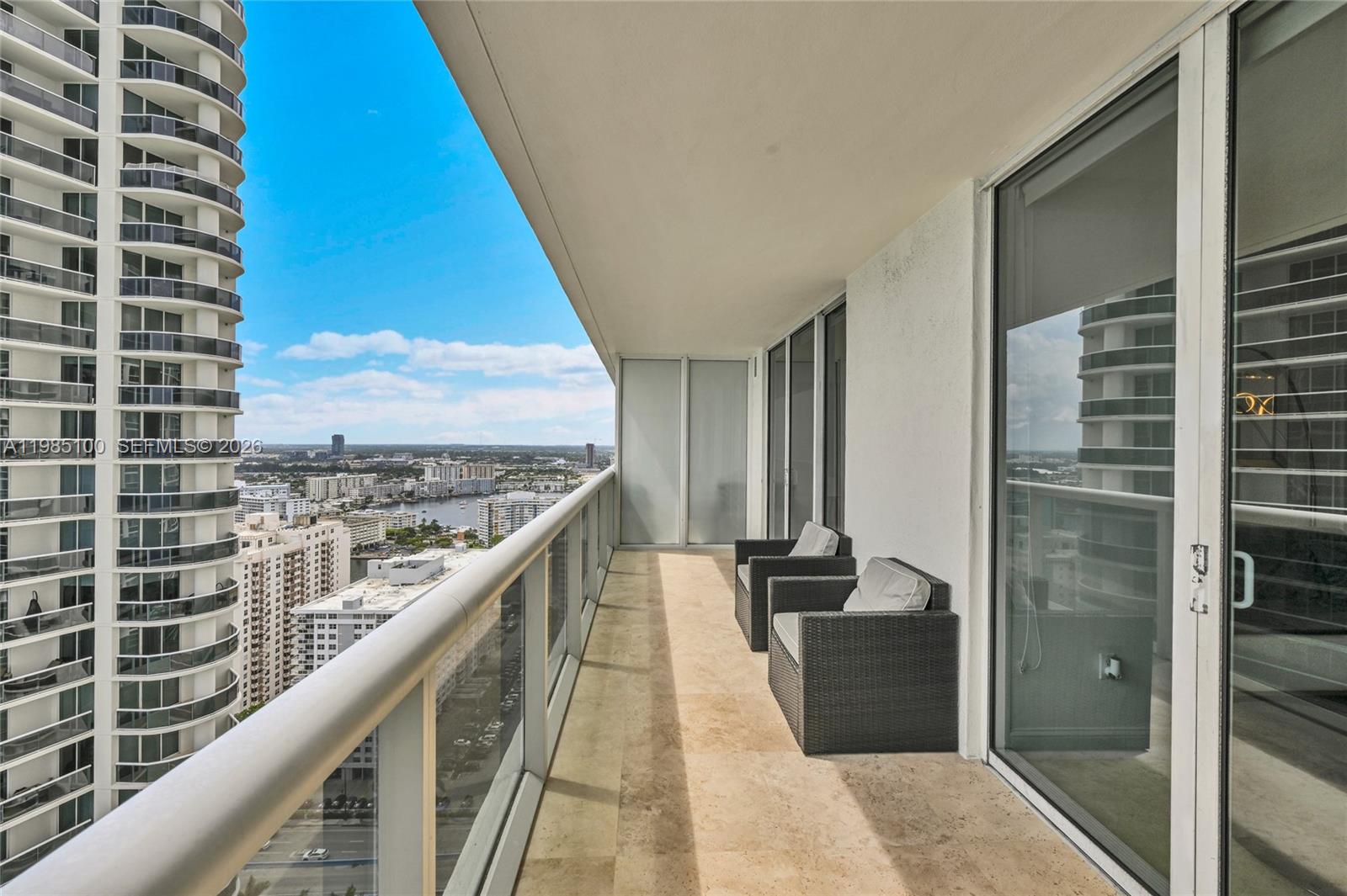 A high-rise balcony features two wicker chairs, tile flooring, and a glass railing overlooking cityscapes — 1830 S Ocean Dr # 3104, Hallandale Beach FL 33009, HALLANDALE BEACH, FL