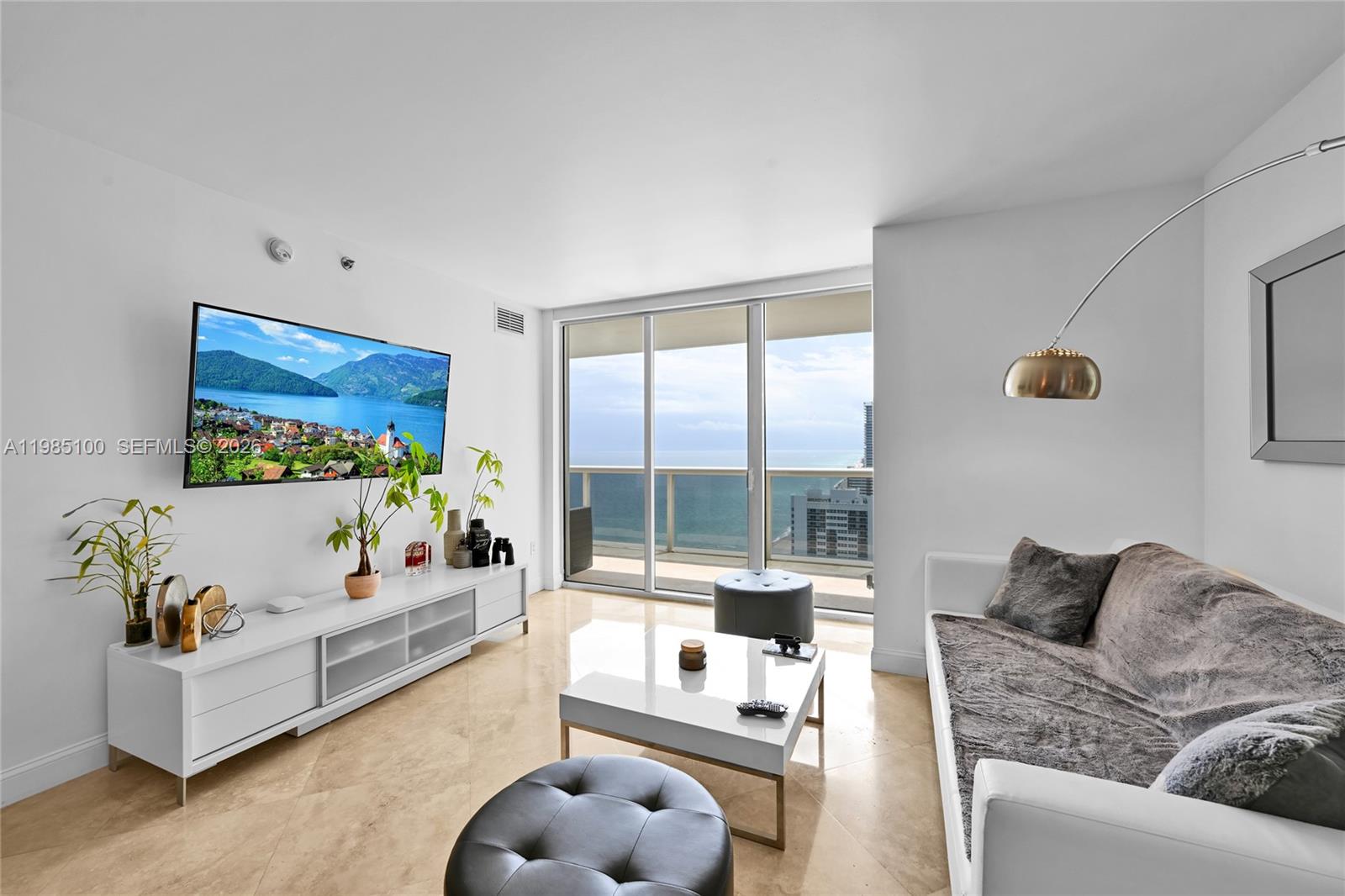 A modern living room features a white media console, a sofa, and a balcony view — 1830 S Ocean Dr # 3104, Hallandale Beach FL 33009, HALLANDALE BEACH, FL