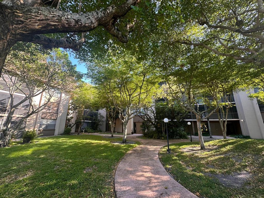 A paved walkway leads through a grassy courtyard toward multi-story apartment buildings shaded by trees — 661 NE 195TH STREET UNIT 402, MIAMI, FL