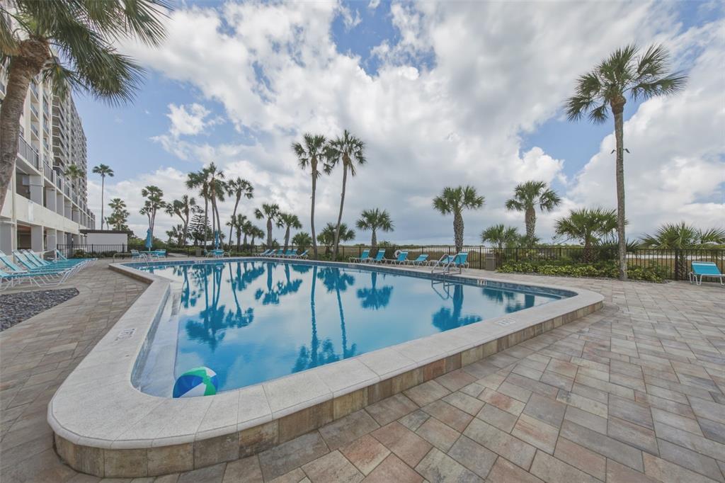 A swimming pool with lounge chairs and palm trees sits beside a multi-story building — 1270 GULF BOULEVARD UNIT 1202, CLEARWATER BEACH, FL