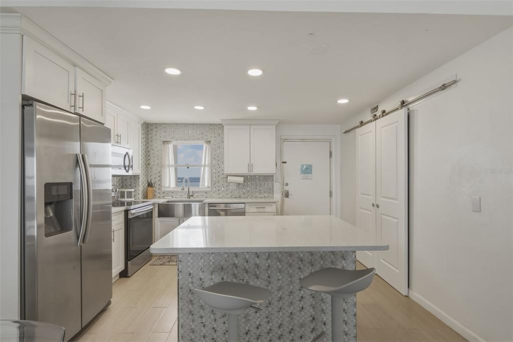A modern kitchen features white cabinetry, a quartz-topped island, stainless steel appliances, and mosaic tiling — 1270 GULF BOULEVARD UNIT 1202, CLEARWATER BEACH, FL