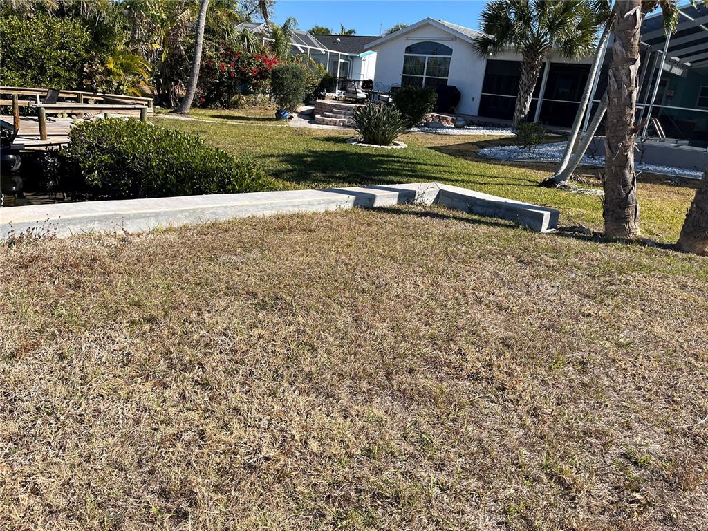 A grassy backyard features a concrete retaining wall, landscaping, and a house in the background — 19262 PINE BLUFF COURT, PORT CHARLOTTE, FL