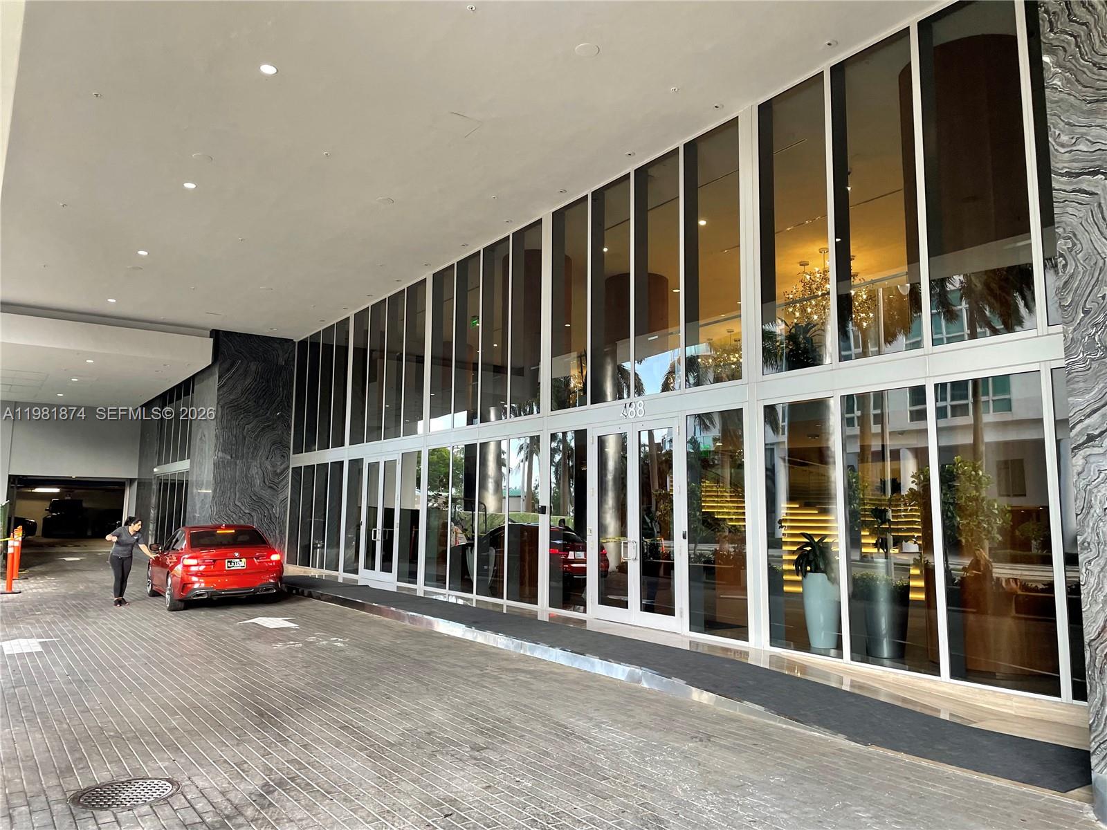 A red car is parked under the covered entrance of a modern glass-fronted building — 488 NE 18th St # 1000, Miami FL 33132, MIAMI, FL
