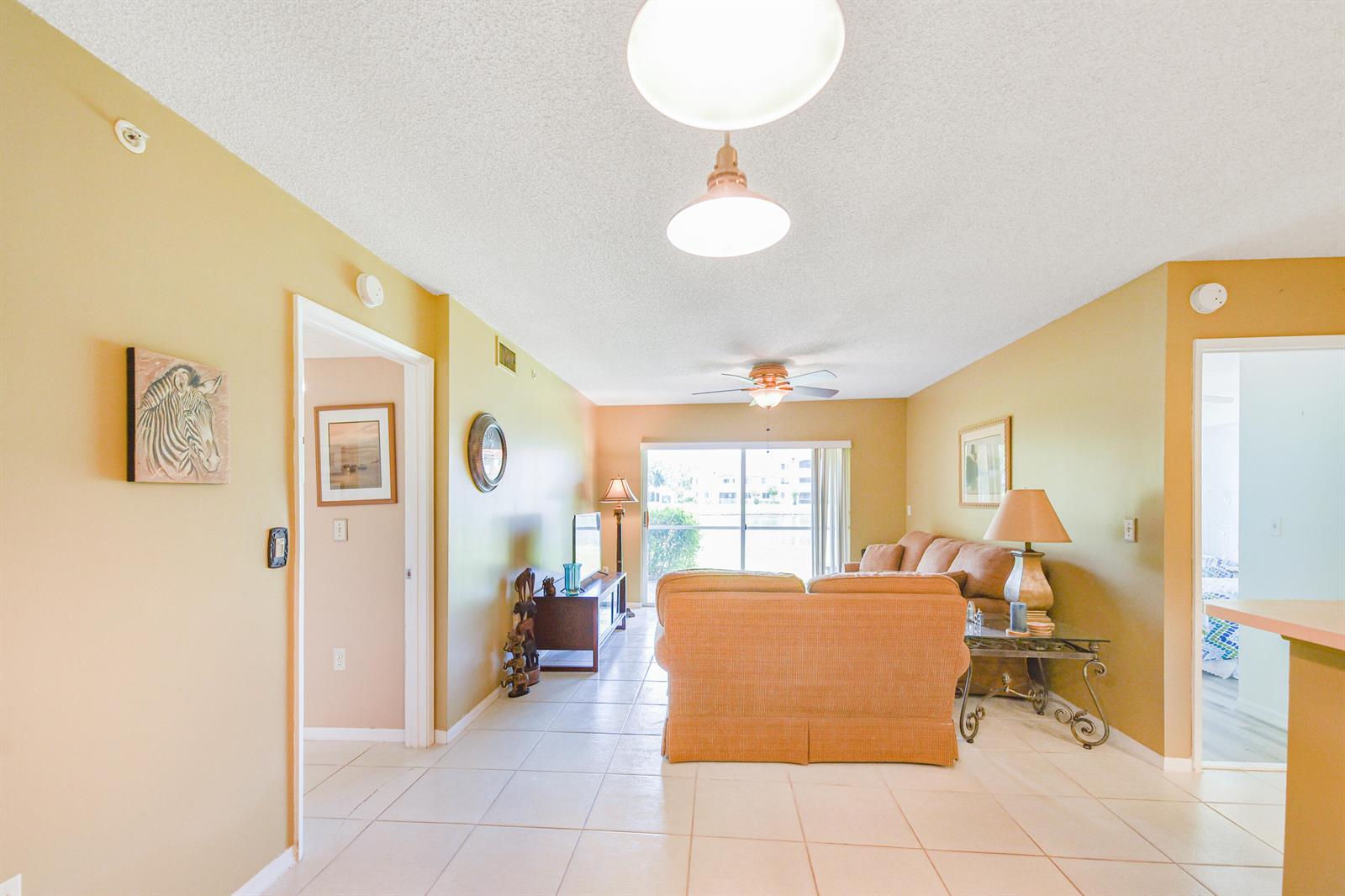 A living room with tan walls, tile flooring, a sofa, and a sliding glass door — 1590 S 42nd Circle # 106, Vero Beach FL 32967, VERO BEACH, FL