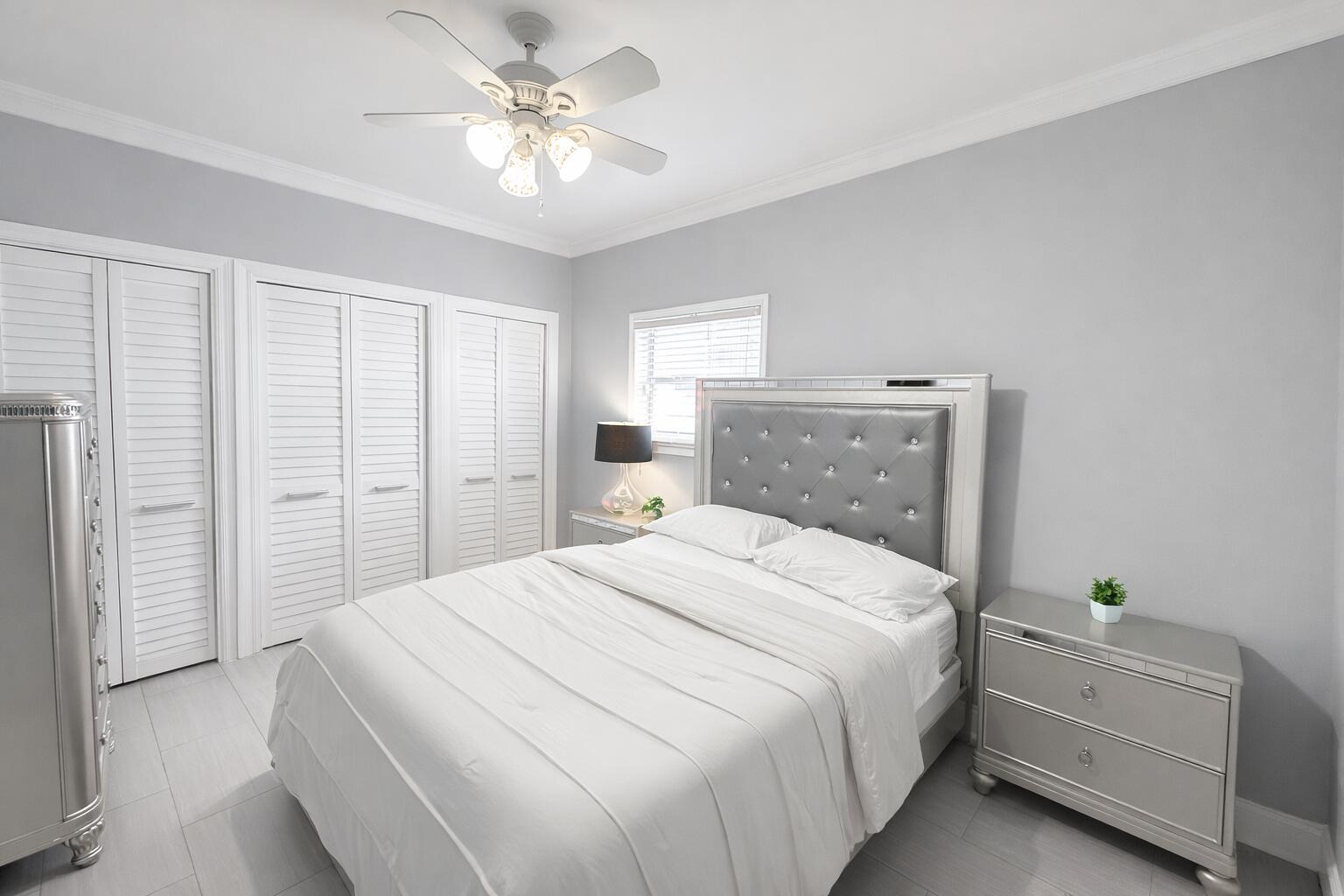 A bedroom features a tufted silver headboard, white bedding, louvered closet doors, and light flooring — 1827 Len Drive, North Palm Beach FL 33408, NORTH PALM BEACH, FL
