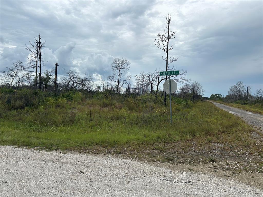A gravel road intersection features a street sign labeled Marabella Rd amidst a grassy, wooded lot — 3520 MARIANA AVENUE, LAKE PLACID, FL