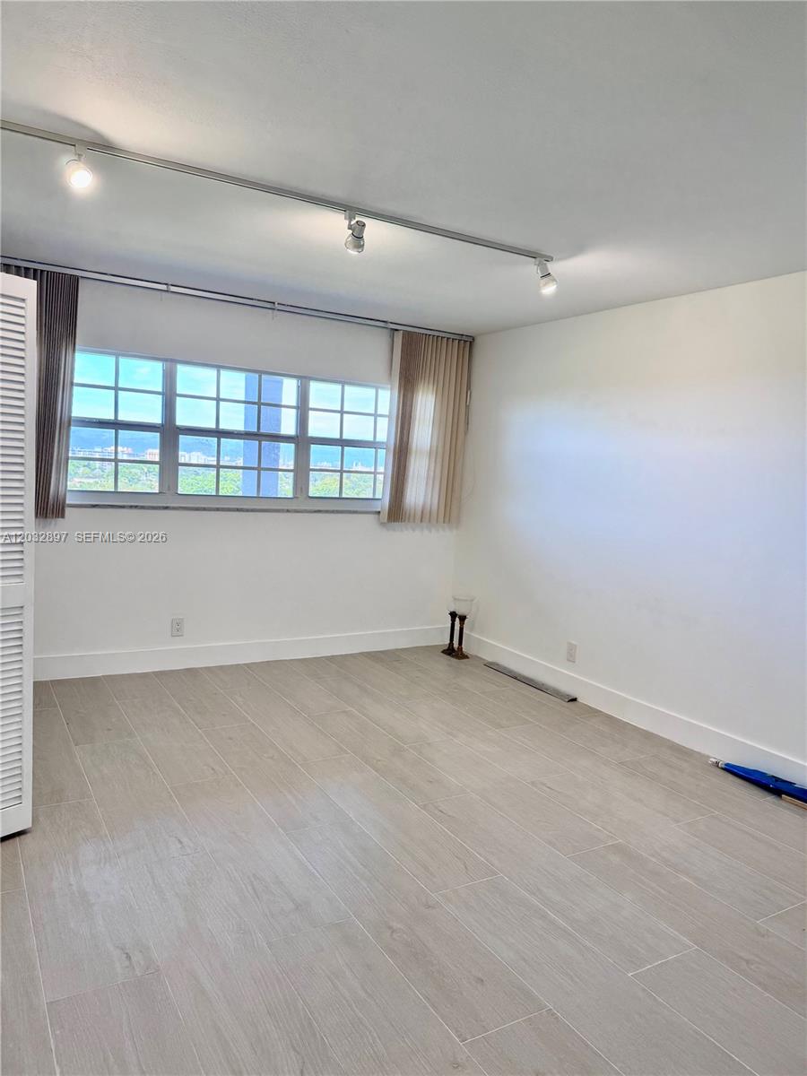 An empty room features light wood-look tile flooring, a track lighting system, and a window — 575 Oaks Ln # 1008, Pompano Beach FL 33069, POMPANO BEACH, FL