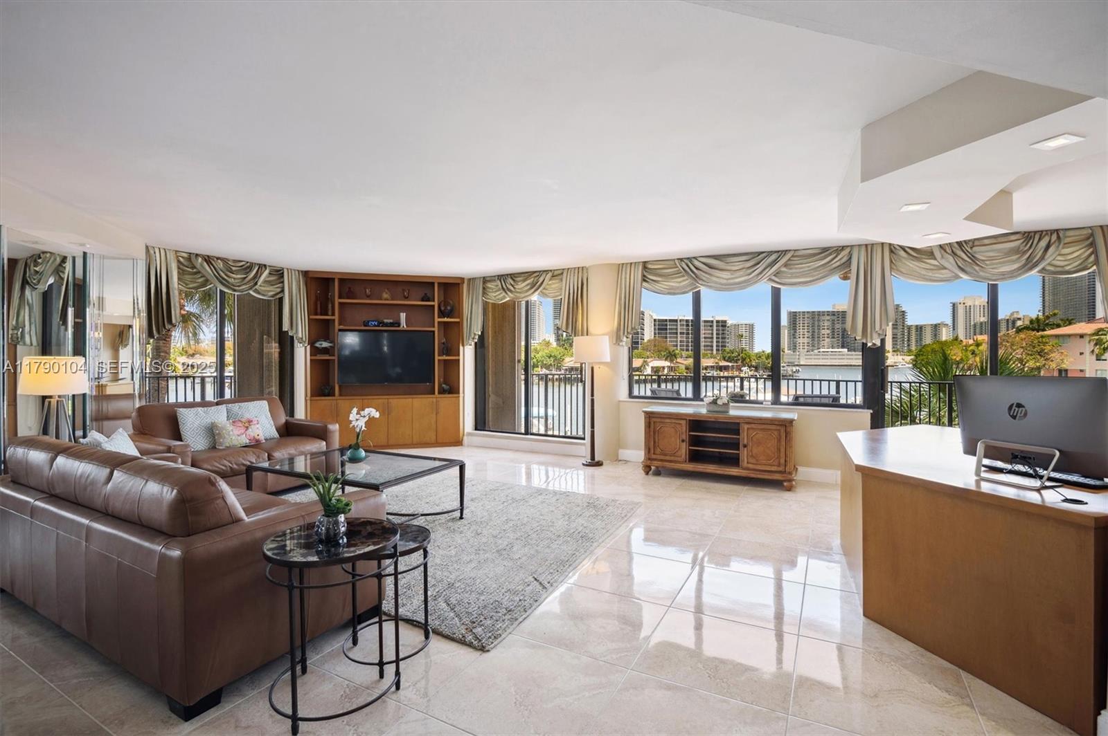 A living room with brown leather sofas, tile flooring, and large windows overlooking the water — 300 Three Islands Blvd # 601, Hallandale Beach FL 33009, HALLANDALE BEACH, FL