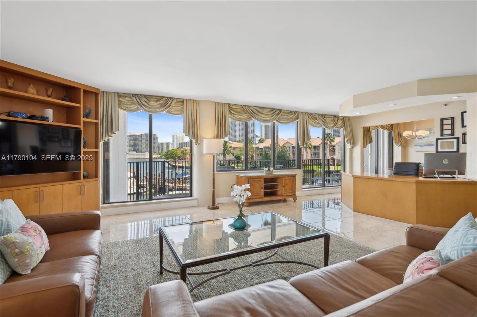 A living room features leather sofas, a glass coffee table, built-in shelving, and waterfront views — 300 Three Islands Blvd # 601, Hallandale Beach FL 33009, HALLANDALE BEACH, FL