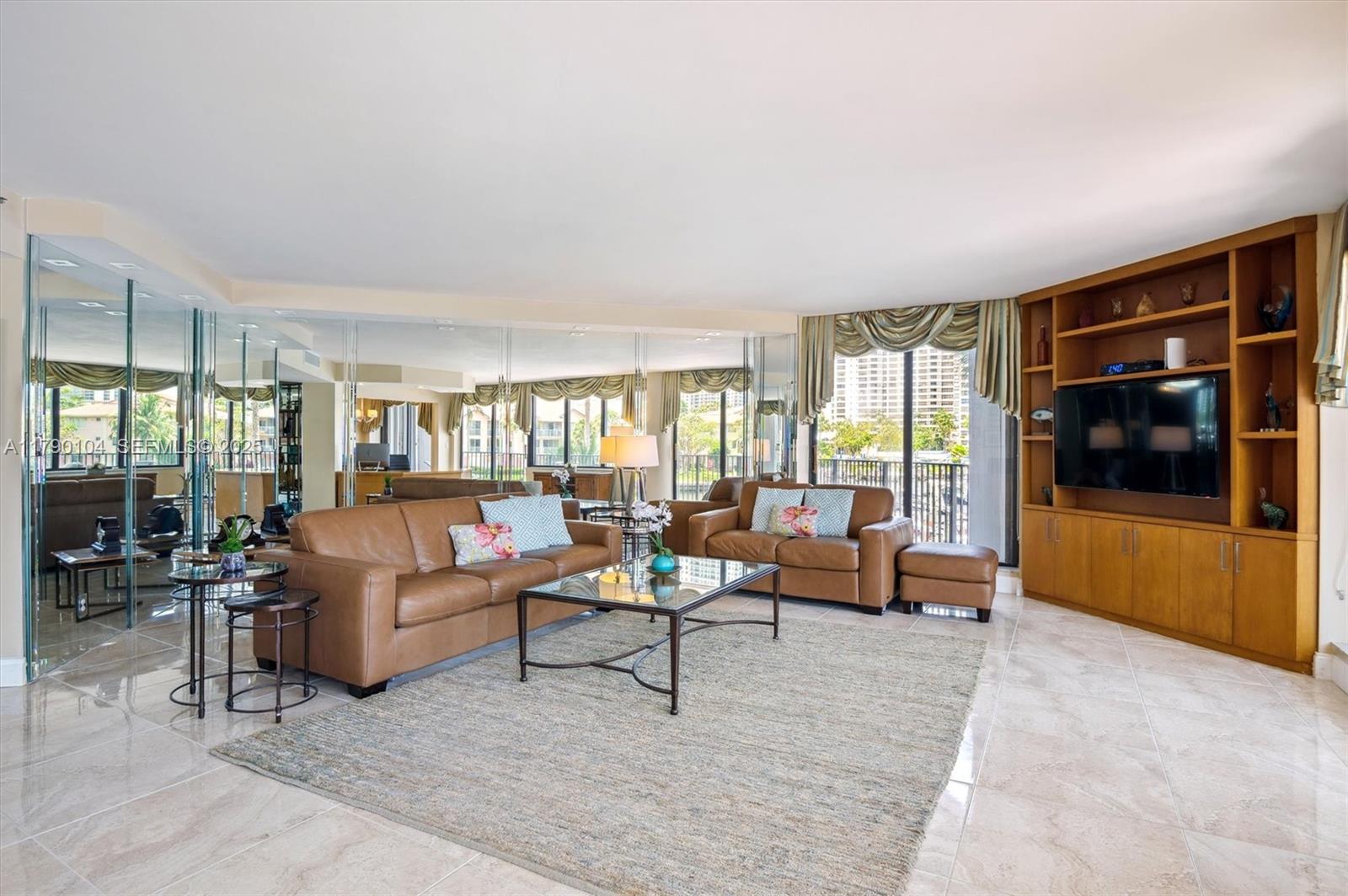A living room features leather sofas, a glass coffee table, and a built-in wooden entertainment center — 300 Three Islands Blvd # 601, Hallandale Beach FL 33009, HALLANDALE BEACH, FL