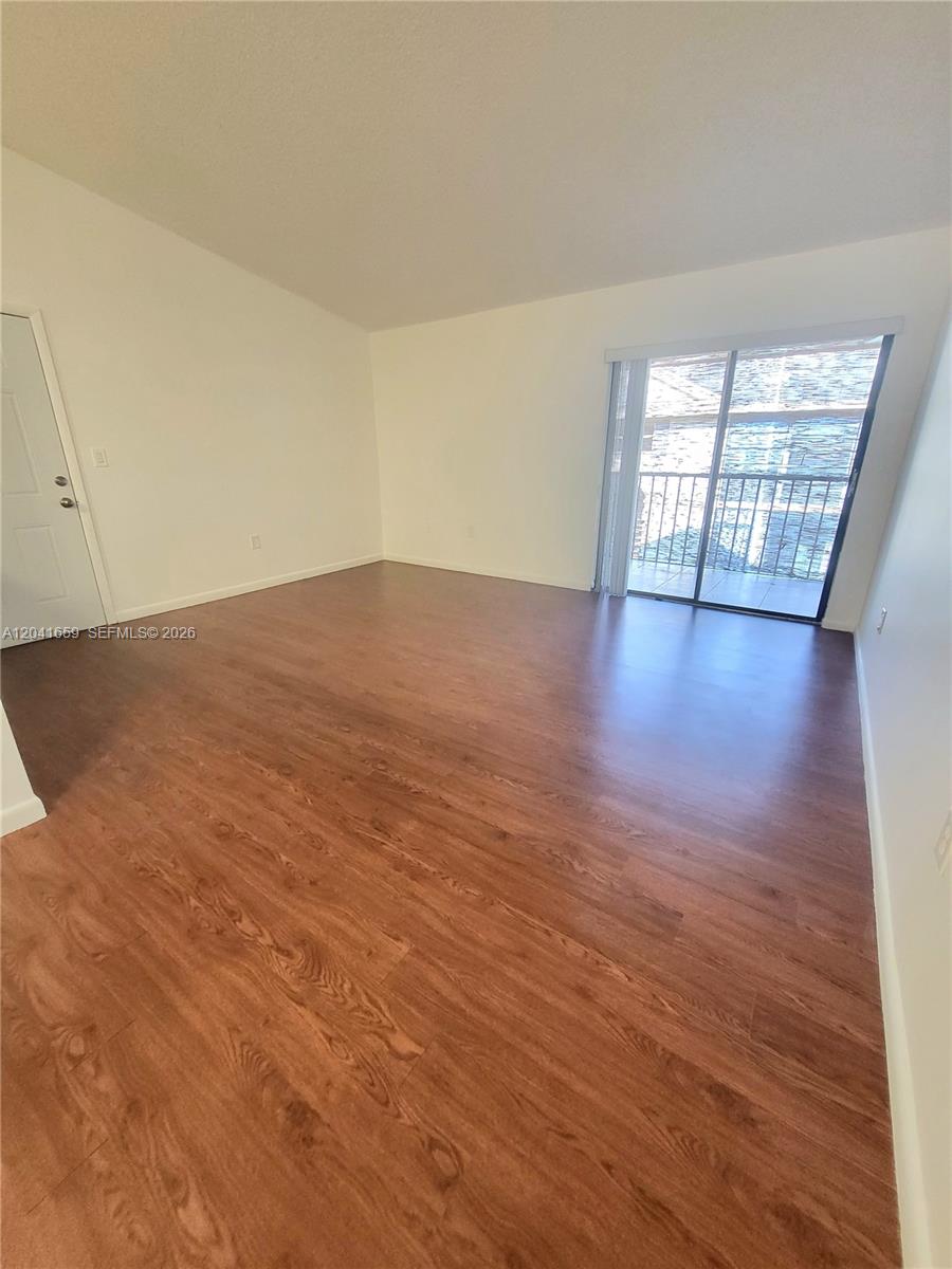 An empty room features wood-look flooring, white walls, and a sliding glass door to a balcony — 18845 NW 62nd Ave # 207, Hialeah FL 33015, HIALEAH, FL
