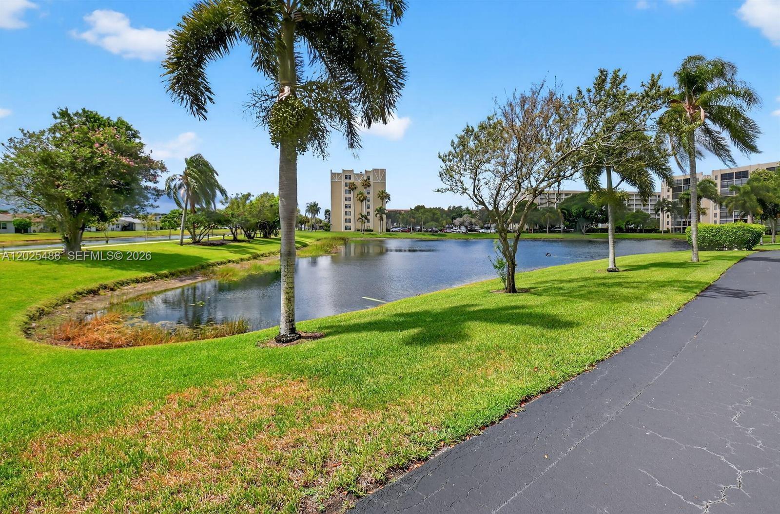 A paved path runs alongside a pond with palm trees and a distant apartment building — 7270 Ashford Pl # 308, Delray Beach FL 33446, DELRAY BEACH, FL