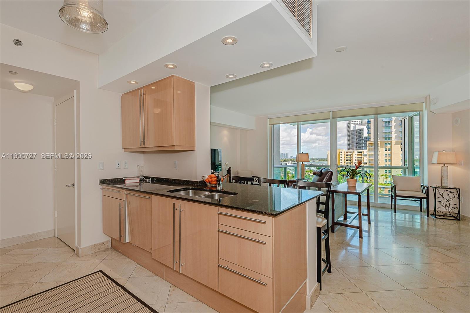 A modern kitchen with light wood cabinets and dark countertops overlooks a bright living area — 1000 S Pointe dr # 906, Miami Beach FL 33139, MIAMI BEACH, FL