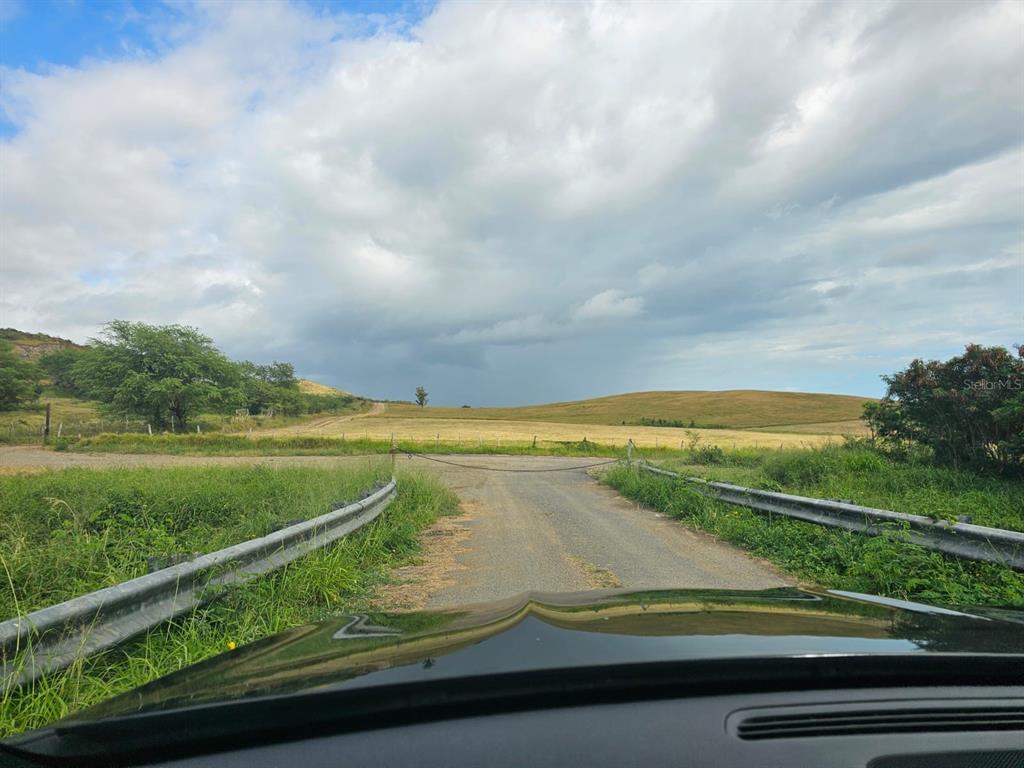 A gravel road leads to a gated entrance overlooking a grassy field under a cloudy sky — BO. LLANOS COSTA SOLAR 7, CABO ROJO, PR