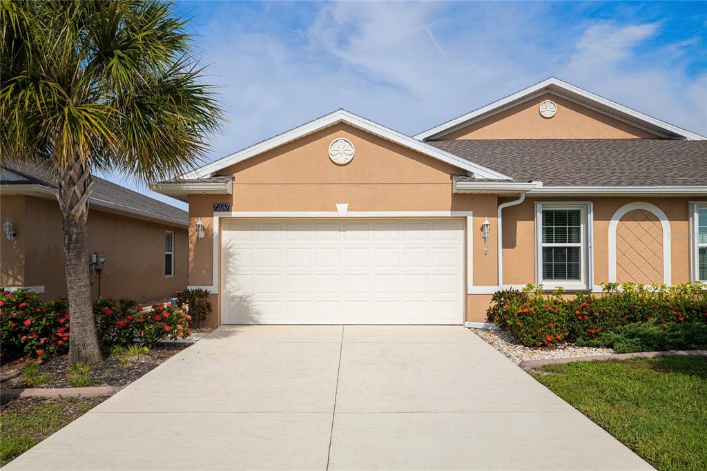 A tan house exterior features a two-car garage, a palm tree, and landscaped bushes — 7097 W LENOX CIRCLE, PUNTA GORDA, FL