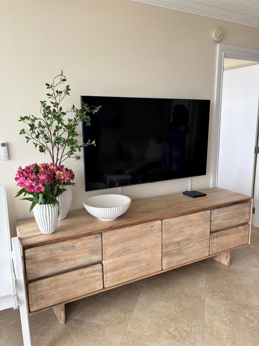 A wall-mounted television sits above a light wood media console with floral arrangements and decor — 531 N Ocean Boulevard # 1107, Pompano Beach FL 33062, POMPANO BEACH, FL