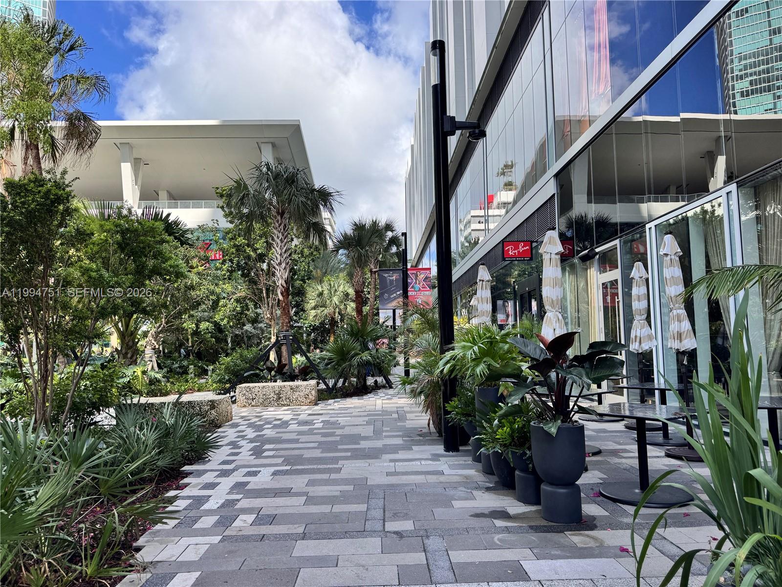 An outdoor pedestrian walkway features stone pavers, potted plants, and modern glass-fronted retail buildings — 159 NE 6 # 4508, Miami FL 33132, MIAMI, FL
