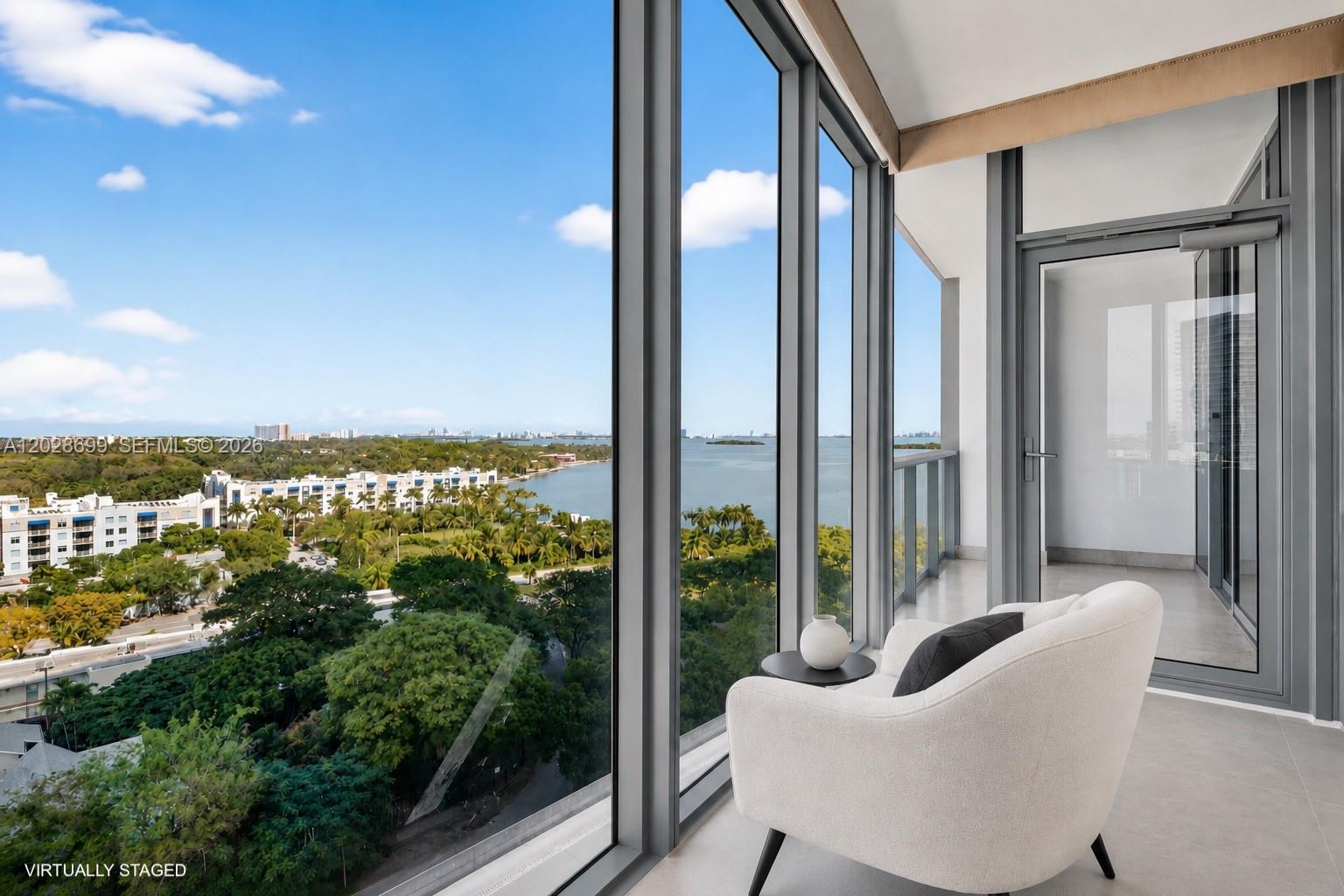A modern balcony features floor-to-ceiling windows, a white armchair, and a view of the bay — 601 NE 36th St # 1101, Miami FL 33137, MIAMI, FL