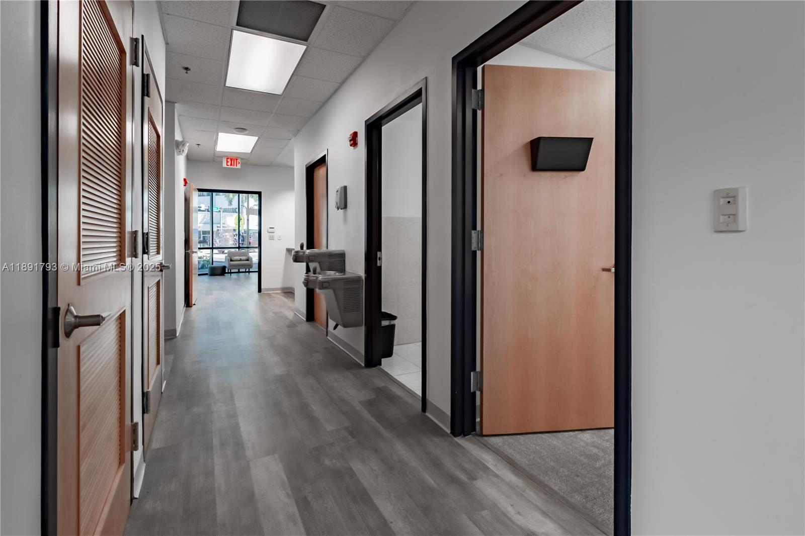 A hallway features wood-look flooring, several wooden doors, a water fountain, and an exit sign — 410 W 41st St # 103, Miami Beach FL 33140, MIAMI BEACH, FL