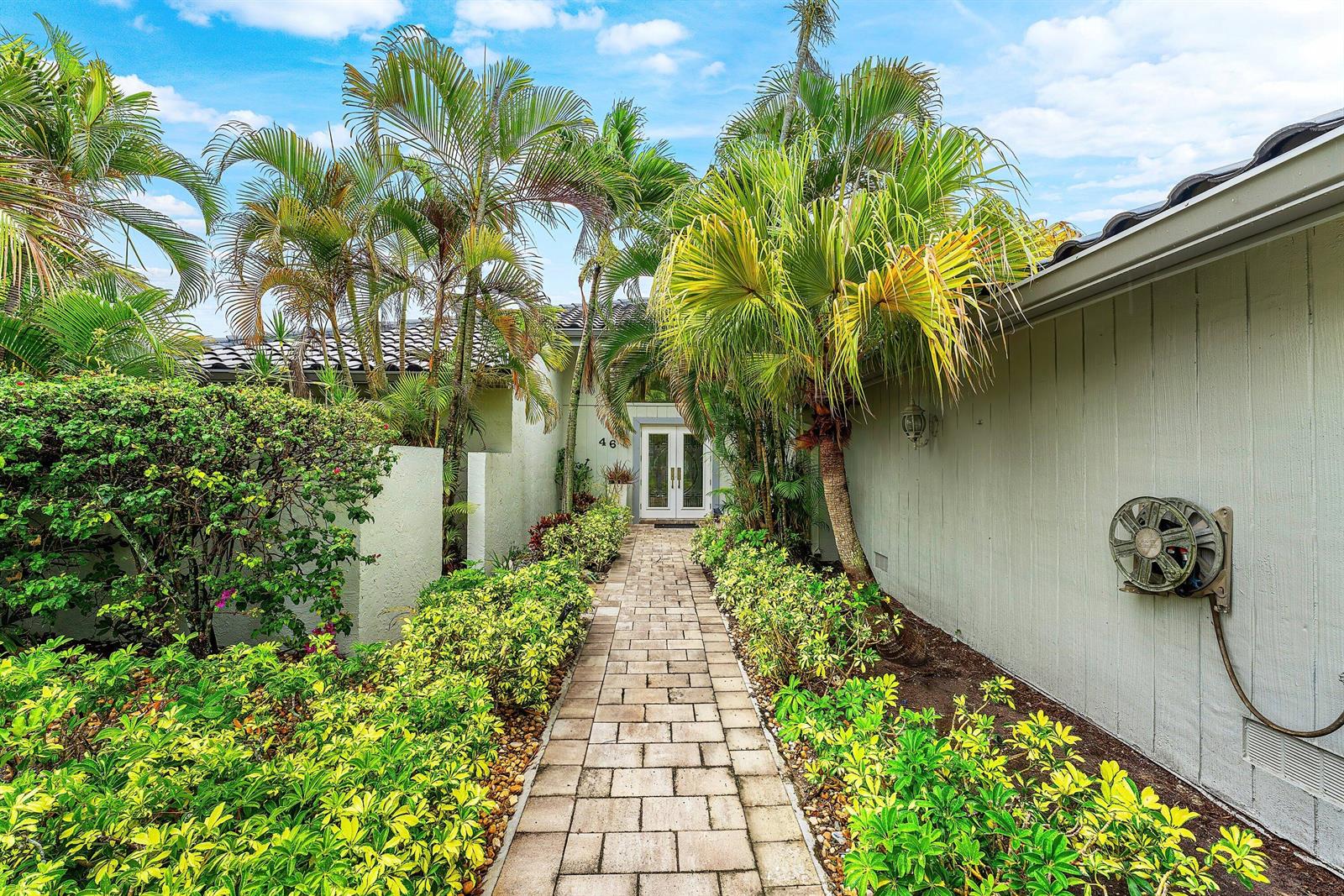 A stone walkway leads to a white front door surrounded by palm trees and landscaping — 46 Woods Lane, Boynton Beach FL 33436, BOYNTON BEACH, FL